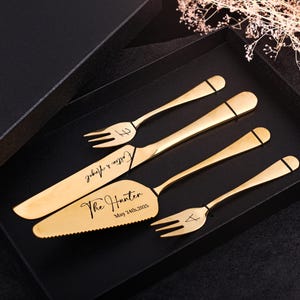 May include: A gold-colored cake serving set in a black box. The set includes a cake knife, server, and two forks. The knife and server are engraved with names and a date. The set is displayed on a black surface.