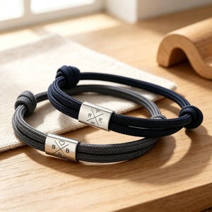 May include: Two rope bracelets with silver metal clasps. One is navy and grey, the other grey. The clasps have engraved symbols and letters. The bracelets are displayed on a wooden surface.