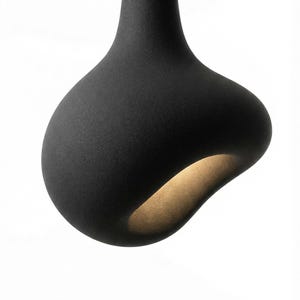 Puede incluir: Lámpara colgante negra moderna con una forma orgánica única. La luz tiene una superficie texturizada y un brillo cálido y acogedor que emana del interior. La lámpara cuelga de un fino cable negro sobre un fondo blanco.