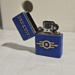 Könnte beinhalten: Blaues Metallfeuerzeug mit dem Text "VAULT-TEC" auf dem Deckel. Das Feuerzeug hat ein goldfarbenes Emblem auf der Vorderseite. Das Feuerzeug ist geöffnet und zeigt die inneren Komponenten.