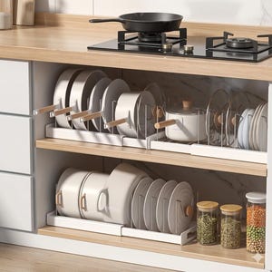 Puede incluir: Un mueble de cocina con utensilios de cocina y vajilla organizados. El mueble presenta ollas, sartenes y platos blancos, con asas y tapas de madera. Se ven frascos de vidrio con especias en el estante inferior, y una sartén negra en la estufa.