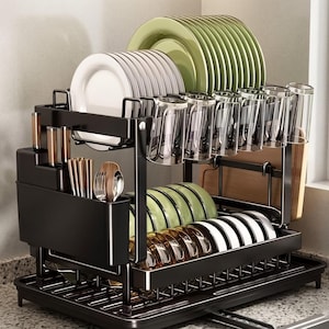 May include: A two-tiered black dish rack with multiple compartments for plates, glasses, and utensils. The rack holds white, green, and clear dishes, along with silverware and a wooden cutting board. The rack sits on a countertop.