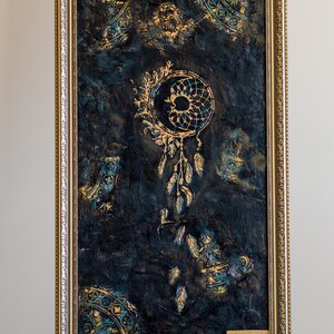 May include: Framed artwork featuring a dreamcatcher design in gold and teal against a dark blue textured background. The artwork is in a gold ornate frame. The dreamcatcher has feathers hanging below.