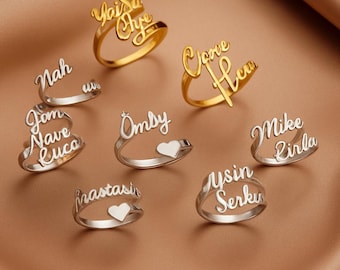 May include: A collection of personalized name rings in gold and silver. The rings feature various names in a cursive font. Some rings have heart-shaped accents. The rings are displayed on a brown surface.