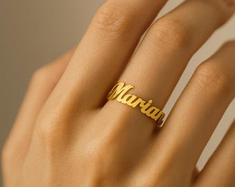 May include: A gold-colored ring spelling out the name "Marian" in a cursive font. The ring is worn on a finger, showcasing the elegant design and personalized touch. The ring is a piece of jewelry.
