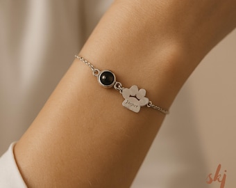 May include: A silver bracelet featuring a dark gemstone and a paw print charm. The paw print charm is engraved with the name "Jasper". The bracelet is delicate and has a simple chain design.