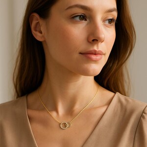 May include: A gold necklace with two intertwined circles, each embellished with small, sparkling details. The necklace hangs from a fine chain. The person wearing the necklace is wearing a tan top.