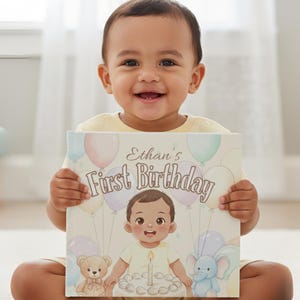 May include: A baby smiles, holding a book that reads "Ethan's First Birthday." The cover shows a cartoon baby, balloons, a teddy bear, and an elephant. The design uses pastel colours.