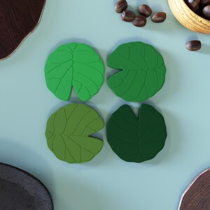 May include: Four green lily pad-shaped coasters. Each coaster has a detailed leaf vein design. The coasters are arranged in a square pattern on a light blue surface. A wooden bowl and scattered nuts are in the background.