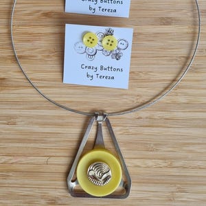 Può includere: Un set di collana e orecchini con dettagli di bottoni gialli. La collana ha un ciondolo color argento con un bottone giallo e un elemento decorativo argentato. Gli orecchini sono borchie con bottoni gialli. Il testo "Crazy Buttons by Tereza" è sulle carte.