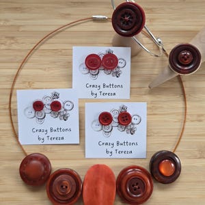 Könnte beinhalten: Eine Kollektion handgefertigter Schmuckstücke und Accessoires mit großen, dekorativen Knöpfen in Rot- und Brauntönen. Das Set umfasst eine Halskette, ein Armband und einen Ring, sowie Karten mit der Aufschrift "Crazy Buttons by Tereza."