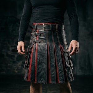 Op de afbeelding: Een zwart en rood leren kilt met een brede tailleband met metalen gesp, twee zijzakken met riempjes en verticale plooien. Het kledingstuk is geconstrueerd met afwisselende zwarte en rode leren panelen en is voorzien van metalen hardware.