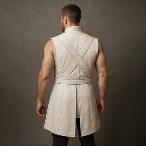 May include: A sleeveless, off-white leather tunic with a high collar and a belted waist. The tunic features a cross-stitch design on the back and a split hem. The garment is worn over black pants.