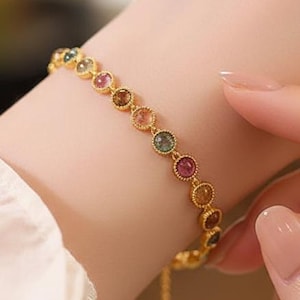 May include: A gold-colored bracelet with a series of colorful gemstones. The gemstones are round and set in individual bezels. The bracelet is on a person's wrist.