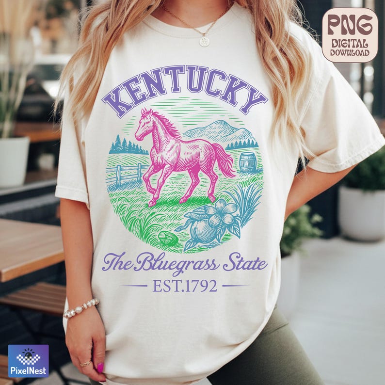 May include: Cream-colored t-shirt featuring a graphic of a pink horse within a circle, with the word "Kentucky" above and "The Bluegrass State" below. The design includes a barrel, flowers, and the text "EST.1792".