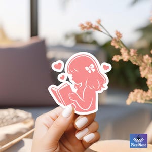 Puede incluir: Una pegatina rosa que representa la silueta de una persona leyendo un libro, con corazones. La pegatina tiene un borde blanco y est&aacute; sujeta por una mano con esmalte de u&ntilde;as blanco. El fondo est&aacute; borroso.