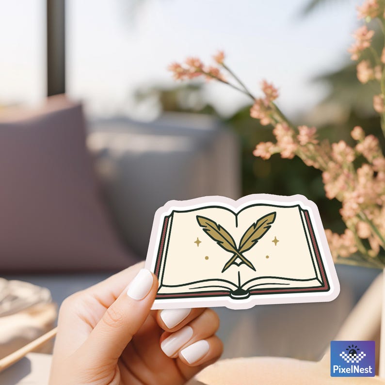 May include: A sticker featuring an open book with two crossed quill pens. The book has a cream-colored interior with gold stars and a dark brown border. The sticker is held by a person with white nail polish.