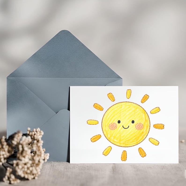 May include: A white greeting card featuring a smiling sun illustration with yellow and orange accents. A blue envelope is behind the card. The card and envelope are on a neutral-colored surface.