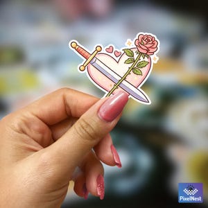 Puede incluir: Una pegatina con un coraz&oacute;n rosa atravesado por una espada y una rosa rosa. La espada tiene una empu&ntilde;adura dorada y la rosa tiene hojas verdes. Peque&ntilde;os corazones rosas y destellos dorados rodean el coraz&oacute;n.