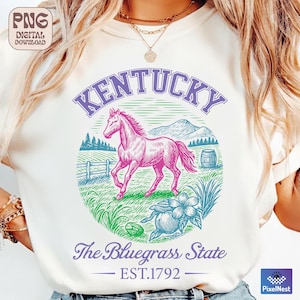 May include: Cream-colored t-shirt featuring a graphic of a pink horse within a circle, with the word "Kentucky" above. Below, it reads "The Bluegrass State" and "EST.1792". The design includes a barrel, flowers, and a fence.