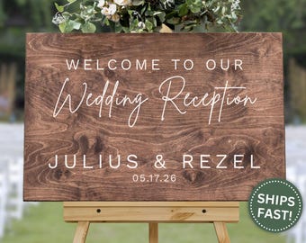 May include: A wooden wedding reception sign with the text "WELCOME TO OUR Wedding Reception" and the names "JULIUS & REZEL" with the date "05.17.26" in white lettering. The sign is displayed on a wooden easel and decorated with flowers.