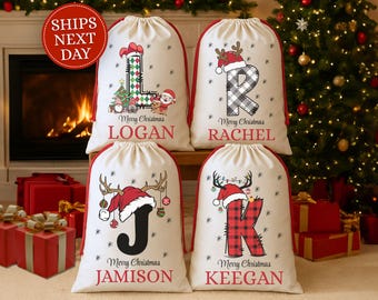 May include: Four cream-colored Christmas gift bags with red trim, each featuring a letter, name, and holiday graphics. The letters are decorated with festive patterns and Santa hats. The bags are personalized with names like Logan, Rachel, Jamison, and Keegan.