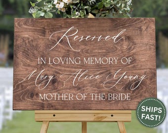 May include: A wooden sign with the words "Reserved" and "In Loving Memory of Mary Alice Young, Mother of the Bride" in white script. The sign is displayed on a wooden easel, with a floral arrangement on top.