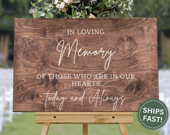 May include: A wooden sign with the words "IN LOVING Memory OF THOSE WHO ARE IN OUR HEARTS Today and Always" in white script. The sign is displayed on a wooden easel, with a floral arrangement at the top.