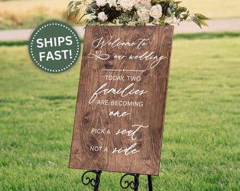 May include: A wooden wedding sign with white calligraphy that reads "Welcome to our wedding" and "Today, two families are becoming one. Pick a seat, not a side." The sign is on a black metal stand.