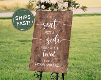 May include: A wooden wedding sign with the text "Pick a seat not a side, you are all loved by the groom and bride" in white script. The sign is on a black metal stand, with a floral arrangement at the top. A green circle with the text "SHIPS FAST!" is in the upper left.