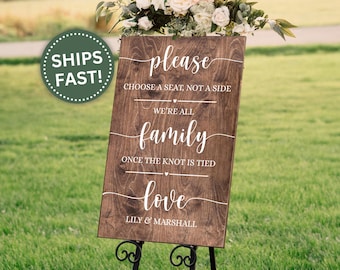 May include: A wooden wedding sign with white calligraphy that reads "please choose a seat, not a side, we're all family, once the knot is tied, love, Lily & Marshall." The sign is on a black metal stand, with a floral arrangement at the top.