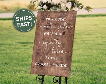 May include: Wooden wedding sign with the text "Pick a seat not a side, you are all equally loved by the groom and bride." The sign is brown with white lettering and is displayed on a black metal stand. A green circle with the words "SHIPS FAST!" is in the upper left corner.