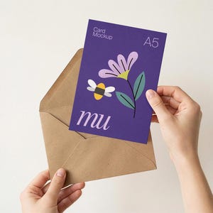 May include: A5 card mockup with a purple background featuring a flower and bee illustration, held in a brown envelope. The card has the text "Card Mockup" and "mu". The envelope is held by two hands.
