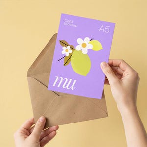 May include: A lilac A5 card mockup with a lemon and flower illustration is held above a brown envelope. The card has the word "mu" printed on it. The background is a solid yellow color.