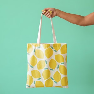 May include: A white tote bag with a repeating pattern of yellow lemons and green leaves. The bag is held by a person against a solid teal background. The bag has white handles.