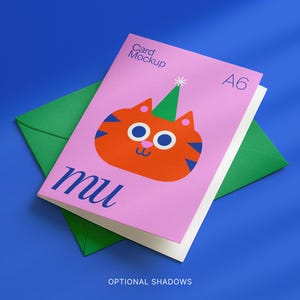 May include: A pink A6 card mockup featuring an orange cartoon cat with a green party hat, on a green envelope. The card has the word "mu" in blue script. The background is a solid blue.