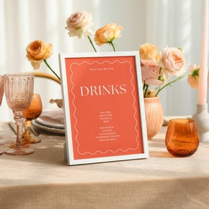 Puede incluir: Una mesa decorada con un letrero enmarcado en blanco que dice "DRINKS" sobre un fondo naranja. La mesa está decorada con cristalería, velas y un jarrón con flores. La escena está bañada en luz suave.