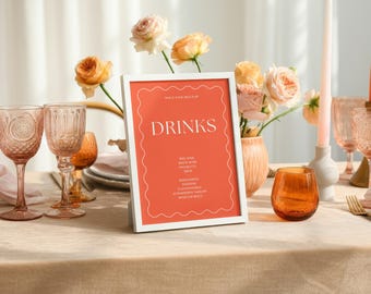 Può includere: Un'apparecchiatura con un cartello con cornice bianca che recita "DRINKS" su sfondo arancione. Il tavolo è apparecchiato con bicchieri, candele e un vaso di fiori. La scena è immersa in una luce soffusa.