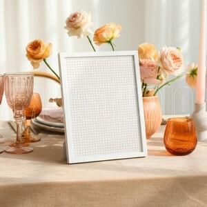 Pu&ograve; includere: Una lavagna bianca con cornice &egrave; appoggiata su un tavolo apparecchiato per un'occasione speciale. Il tavolo &egrave; adornato con bicchieri rosa, candele e un vaso di rose. Un bicchiere arancione &egrave; vicino alla lavagna.