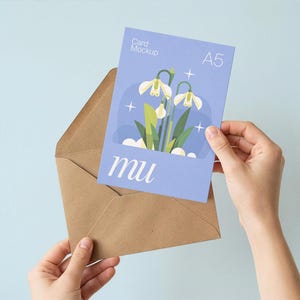 May include: A5 card mockup with a floral illustration and the word "mu" in a light blue envelope. The card features a design of white flowers with green leaves. The background is a light blue.