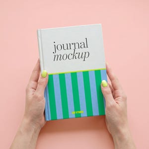 May include: A hardcover journal with a white upper section featuring the black text journal mockup and a lower section with vertical green and light blue stripes. A small yellow logo is printed at the bottom center of the striped area.