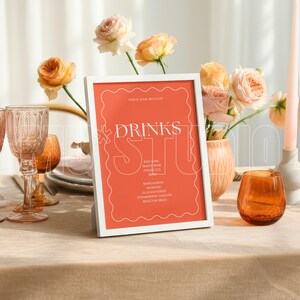 Pu&ograve; includere: Un'apparecchiatura da tavola con un cartello con cornice bianca che recita "DRINKS" in caratteri bianchi. Il cartello &egrave; su sfondo corallo. La tavola &egrave; apparecchiata con bicchieri, fiori e candele.
