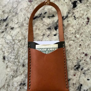 May include: A handmade brown leather wallet with a handle, holding a dollar bill. The wallet has a contrasting black top and is stitched with light brown thread. The handle is secured with a gold-colored rivet.