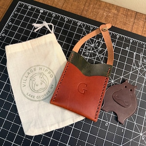 May include: A leather cardholder in shades of brown and green, with a stitched border and a strap. The letter "G" is embossed on the front. A drawstring bag with a hippo logo and a hippo-shaped leather cutout are also present.
