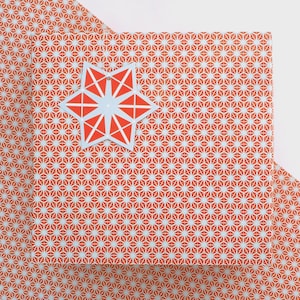 May include: A stack of orange and white patterned wrapping paper with a red and light blue star-shaped ornament. The paper has a repeating geometric design. The ornament is a star shape with a light blue centre and red geometric shapes.