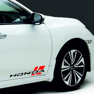Op de afbeelding: Witte auto met zwarte en rode "HONDA" logo-stickers op de zijkant. Het logo heeft de merknaam in zwart en twee rode rechthoeken. De auto heeft zwarte wielen met zilveren accenten.