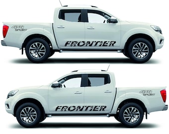 May include: Two white pickup trucks with black lettering that reads "FRONTIER" on the side and "4x4 SPORT" on the rear. The trucks have black wheels and red taillights. The trucks are parked side by side.