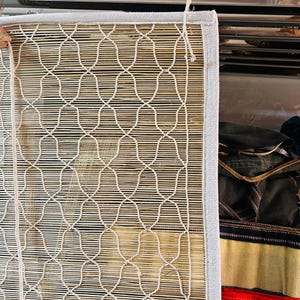May include: A woven bamboo window shade with a white geometric pattern and a white fabric border. The shade is hanging from a metal rod. The bamboo has a natural, light brown color.