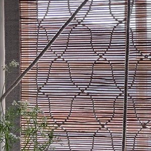 May include: A bamboo window shade with a woven pattern and a rope pull. The shade is a natural brown color and is mounted inside a window frame. A small vase with greenery is in the foreground.