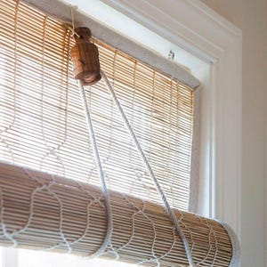 Könnte beinhalten: Ein Bambus-Rollo mit natürlicher Holzoptik. Das Rollo ist aufgerollt und zeigt ein gewebtes Muster. Weiße Kordeln hängen von einer Holzrolle, die zum Heben und Senken des Rollos dient. Der Fensterrahmen ist weiß.
